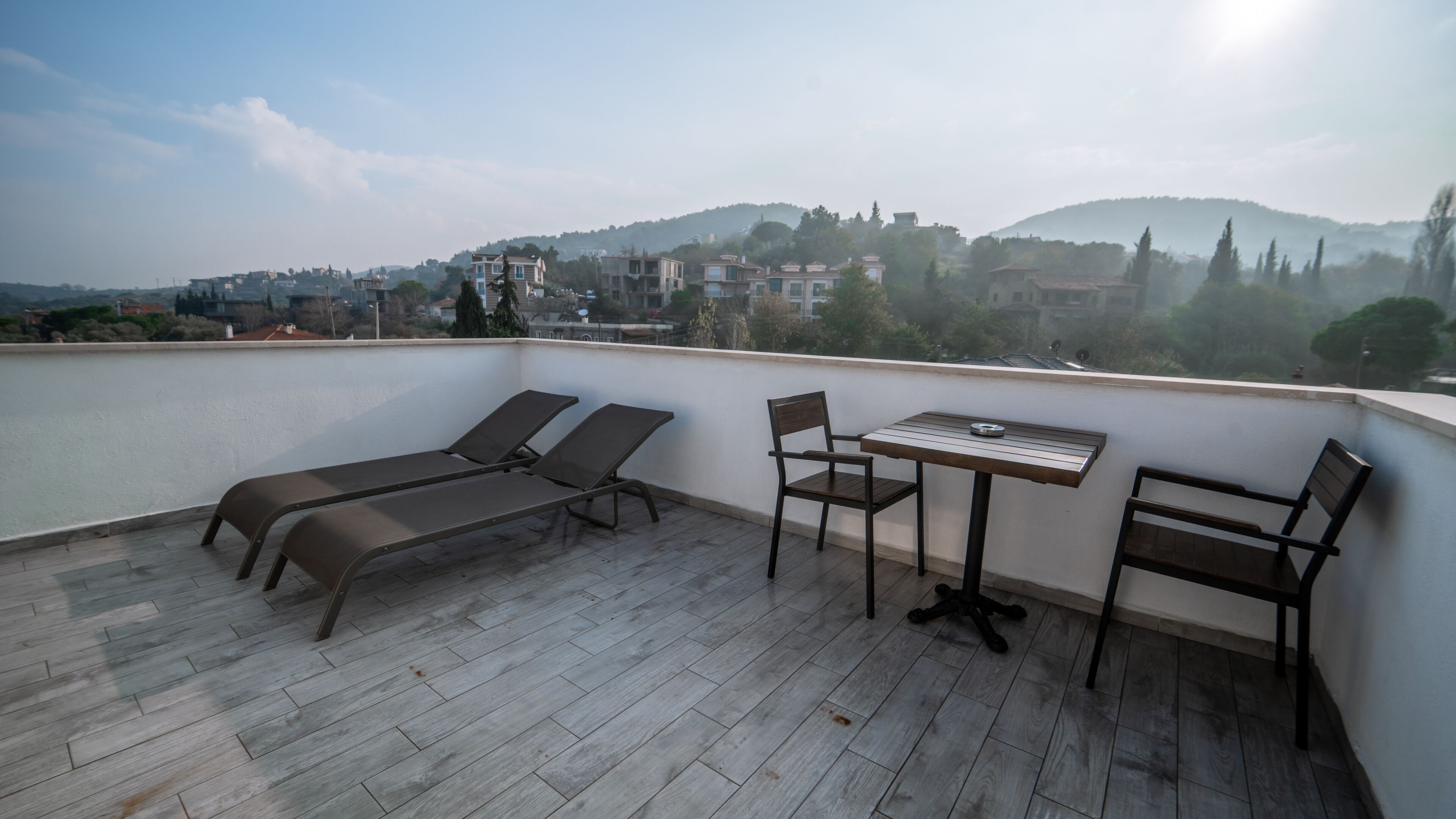 Terraza en la azotea con dos tumbonas, una mesa y dos sillas. La terraza ofrece vistas a un pueblo con edificios y colinas al fondo.