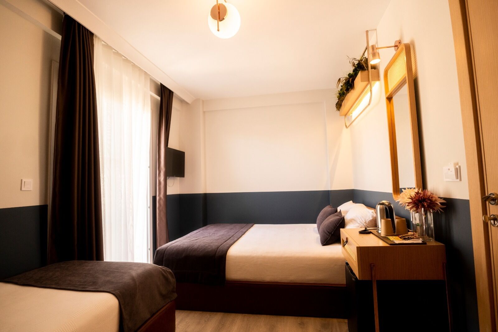 A hotel room with two beds, a bedside table with a lamp, a mirror, and a window with curtains.
