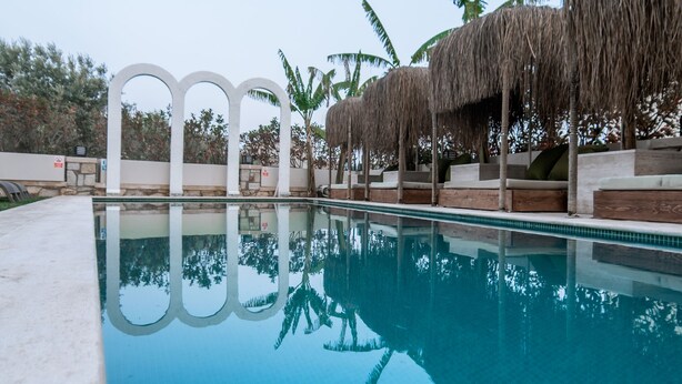 Una piscina con superficie de agua azul cristalina, rodeada de zonas de descanso con sombrillas de paja y palmeras.