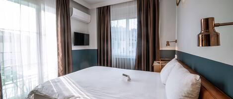 A hotel room with a large bed, a flat-screen TV, an air conditioning unit, a bedside table with a lamp, and a view of the outdoors through the window.