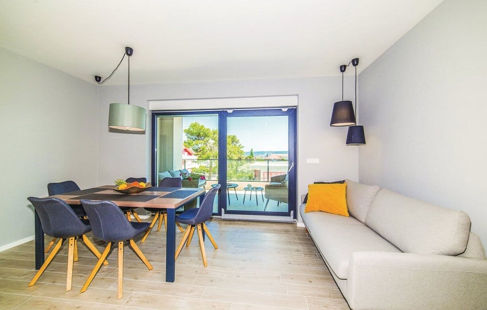 A modern living room with a dining area, large sliding glass doors leading to a balcony, and a sofa with a yellow pillow.