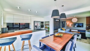 A modern kitchen with white cabinets, a central island, and a dining area with blue chairs and a wooden table. - Luxury Dream House in Kaštel Novi (Kaštel Novi)
