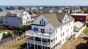 A white two-story house with a balcony, surrounded by other houses, near a body of water. - Glad Tidings - KDH MP 7.5, 320 Yds to Beach, Elevator, Indoor Pool, Hot Tub, Pets OK, Flex Stays (Kill Devil Hills)