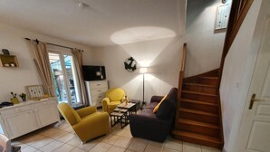 A living room with a sofa, armchair, and a small table with a lamp. There is a staircase to the right and a TV on the wall.