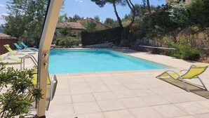 A swimming pool with tiled deck, surrounded by trees and a building with a tiled roof.