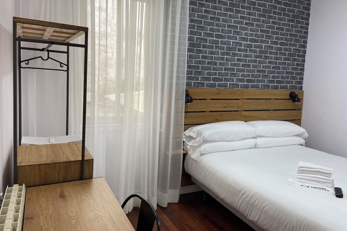 A hotel room with a wooden bed, white bedding, a wooden desk, a chair, a hanging wardrobe, and a brick-patterned wall.