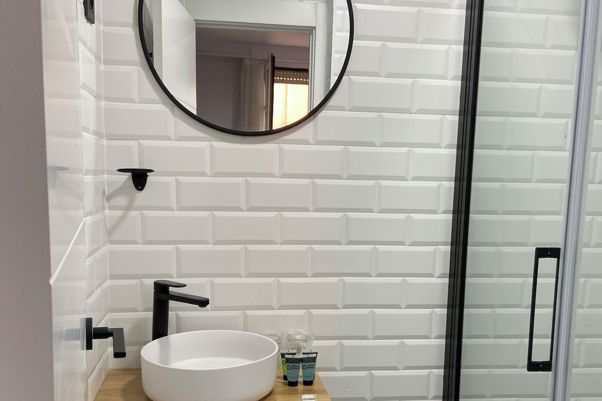 Un baño con lavamanos blanco, grifo negro y un espejo redondo sobre una pared de ladrillo blanco.