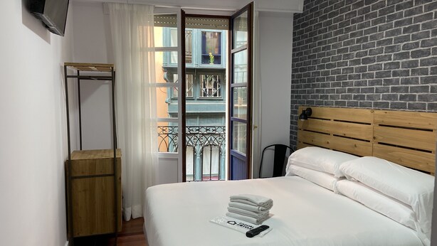 A hotel room with a bed, a wooden headboard, a TV mounted on the wall, a balcony with a view of buildings, and a window with curtains. - Casual Bilbao Mardones (Bilbao)