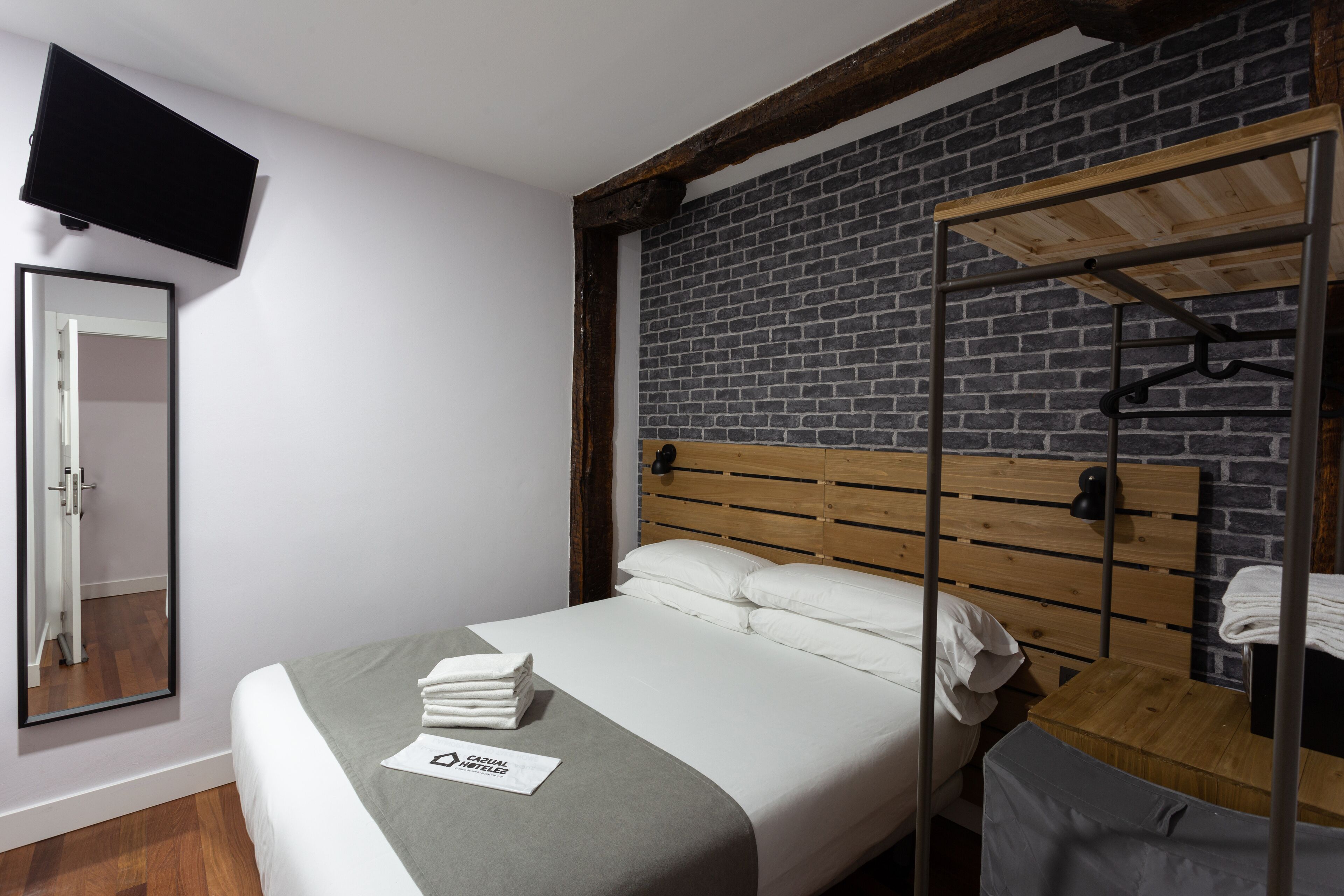 A hotel room with a wooden headboard, a flat-screen TV mounted on the wall, a mirror, a neatly made bed with white linens, and a wooden bench with a towel.
