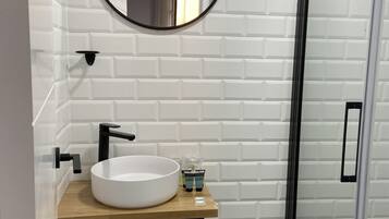 Un baño con lavamanos blanco, espejo y grifo negro. A la derecha, una zona de ducha.