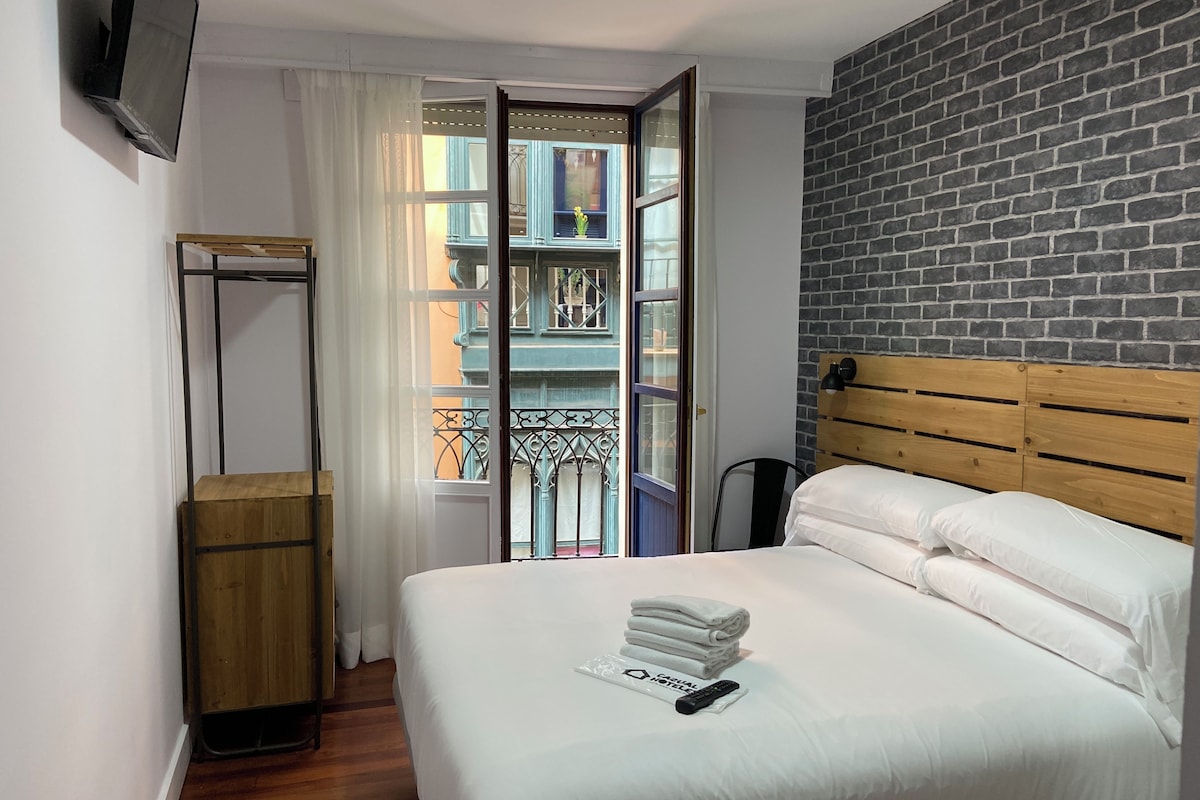 A hotel room with a bed, a wooden headboard, a TV mounted on the wall, a balcony with a view of buildings, and a window with curtains.