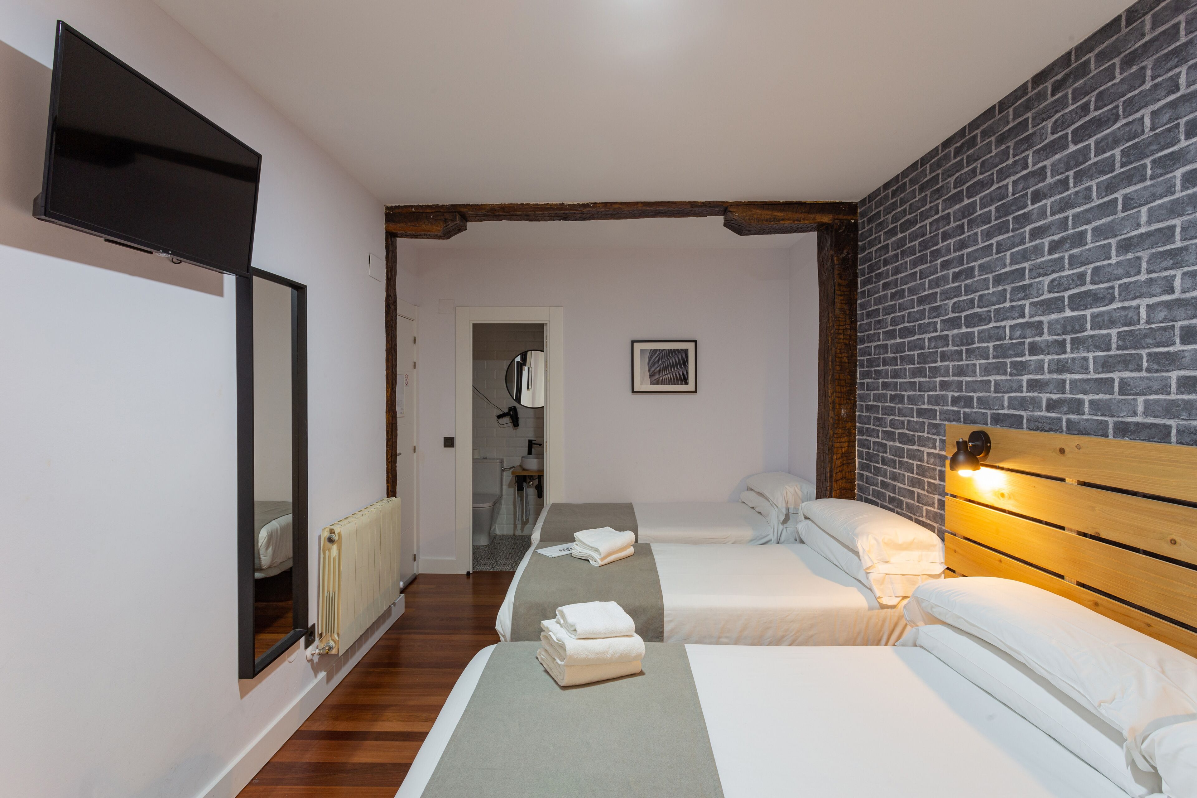 A hotel room with two beds, a large mirror, a brick wall, and a wooden ceiling beam.