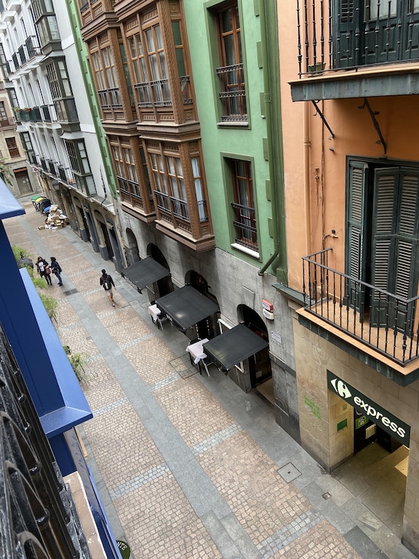 Une vue d’une rue avec des tables à l’extérieur, un bâtiment verdoyant doté de fenêtres décorées, et une enseigne indiquant « service rapide ».