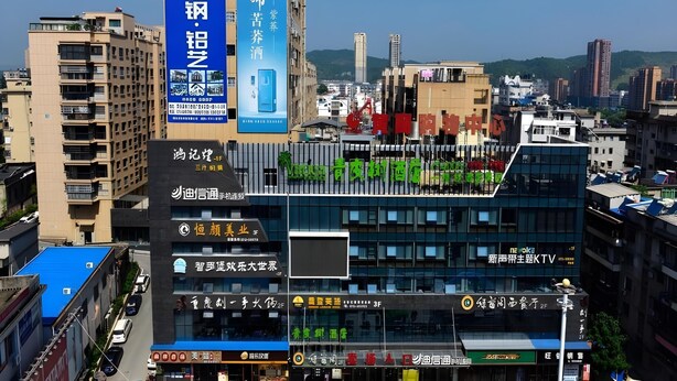 A cityscape with a prominent building featuring a large blue sign with white text, a modern glass facade, and a rooftop garden. - Vatica Huanggang Luotian County City Square Hotel (Huanggang)