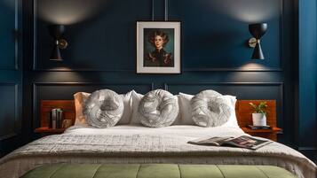 Un dormitorio con pared azul oscuro, un retrato enmarcado, una cama con almohadas estampadas, un banco tapizado de color verde y mesitas de noche con libros y una planta.
