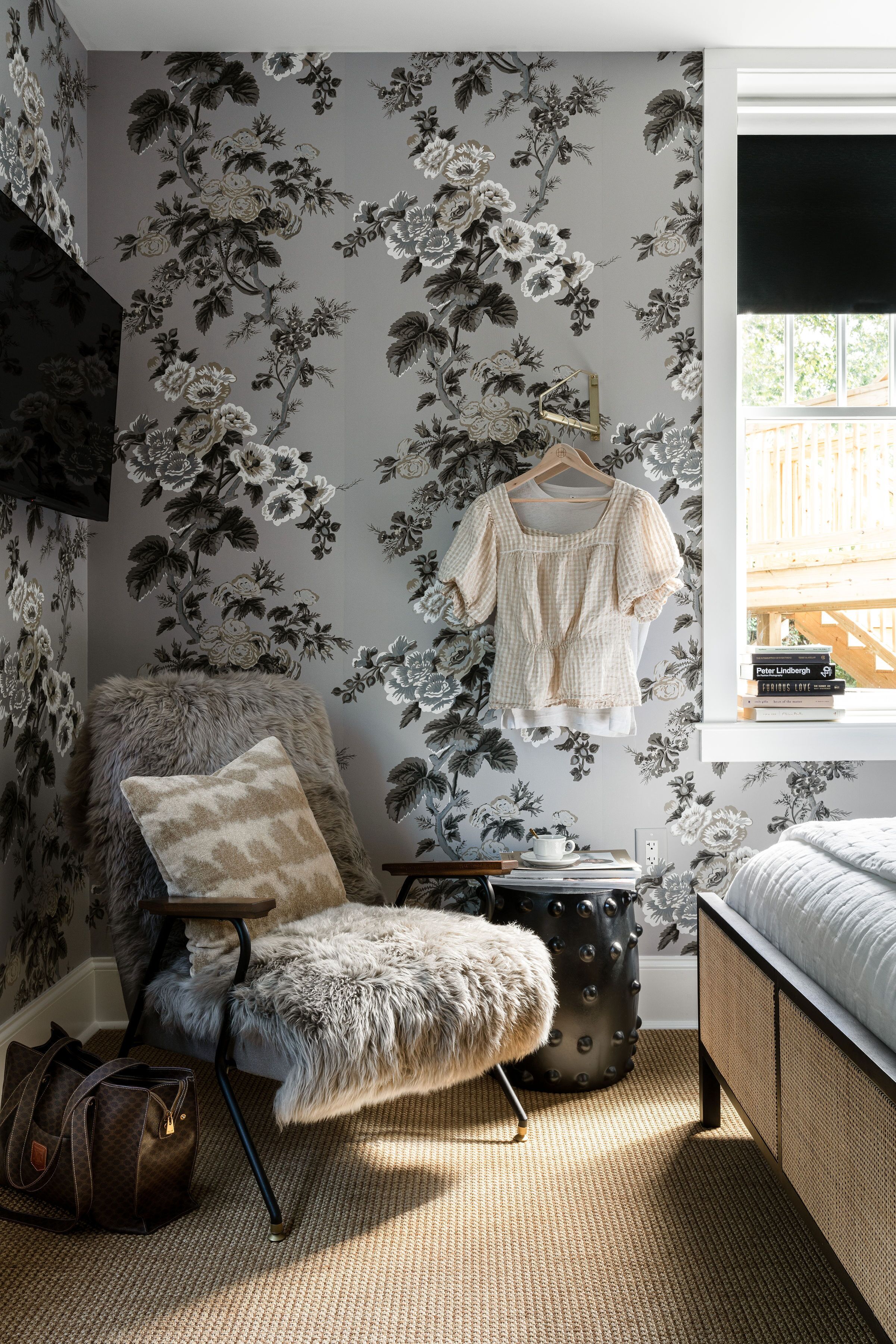 A bedroom with floral wallpaper, a fluffy chair, a hanging garment, a bedside table with books, and a window with a view.