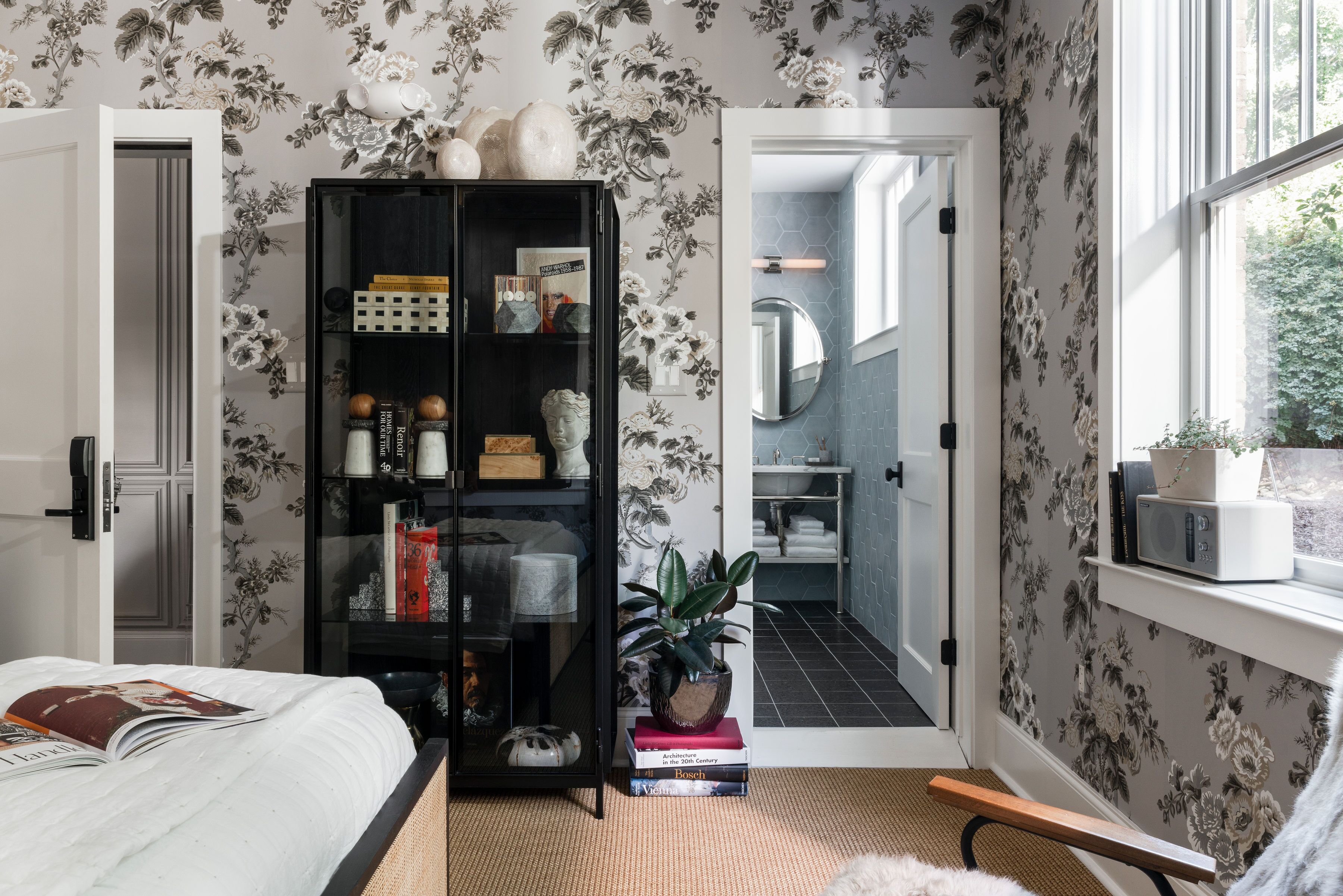 A bedroom with a bed, a black bookcase, a potted plant, and a window with a view of a bathroom.