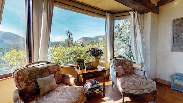 A room with two upholstered chairs, a wooden table, and a view of a mountainous landscape through large windows.