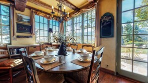 A dining area with a wooden table set for four, surrounded by chairs, with a view of the outdoors through large windows.