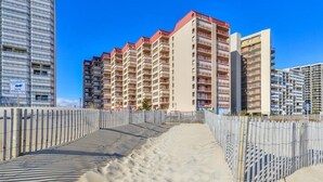 Un hotel frente a la playa con varios edificios altos, un paseo marítimo arenoso y una cerca de madera.