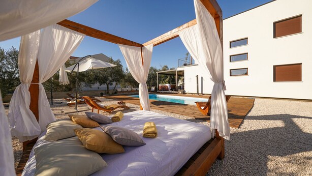 Un dormitorio con cama, almohadas y dosel. Se ve una piscina y sillas de descanso al fondo.