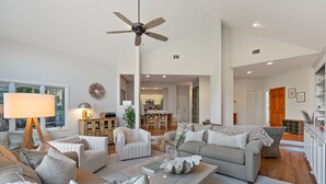 Ein geräumiges Wohnzimmer mit Deckenventilator, einem Essbereich und einer durch eine offene Tür einsehbaren Küche.