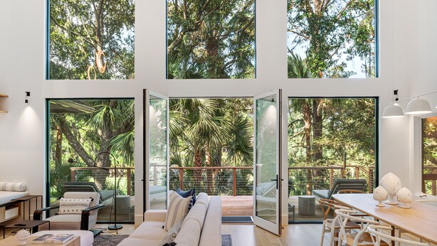 Amplia sala de estar con un sofá grande, una mesa de comedor con sillas y vista a un jardín exuberante a través de grandes ventanales.