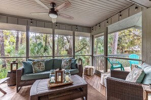 A covered porch with wicker furniture, a ceiling fan, and large windows overlooking a tropical garden.