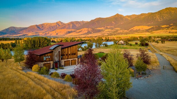 A rustic cabin with a red roof, surrounded by a gravel driveway, trees, and a view of mountains in the background.