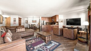 A spacious living room with a large sofa, a coffee table, and a patterned rug. There is a kitchen area with wooden cabinets and a flat-screen TV. - Breckenridge Condo Only a Short Walk to Lifts & Shops w/ 2 Private Balconies! (Breckenridge)
