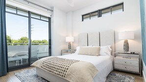 Quarto moderno com uma cama grande, duas mesas de cabeceira, um criado-mudo, porta de vidro que dá para uma varanda e vista para o verde lá fora.
