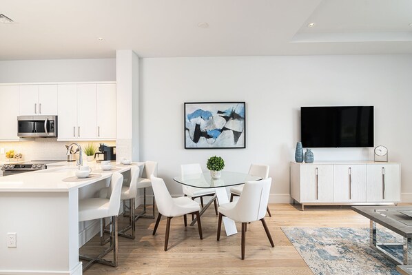A modern kitchen with white cabinetry, a central island with seating, a dining table, and a flat-screen TV mounted on the wall.