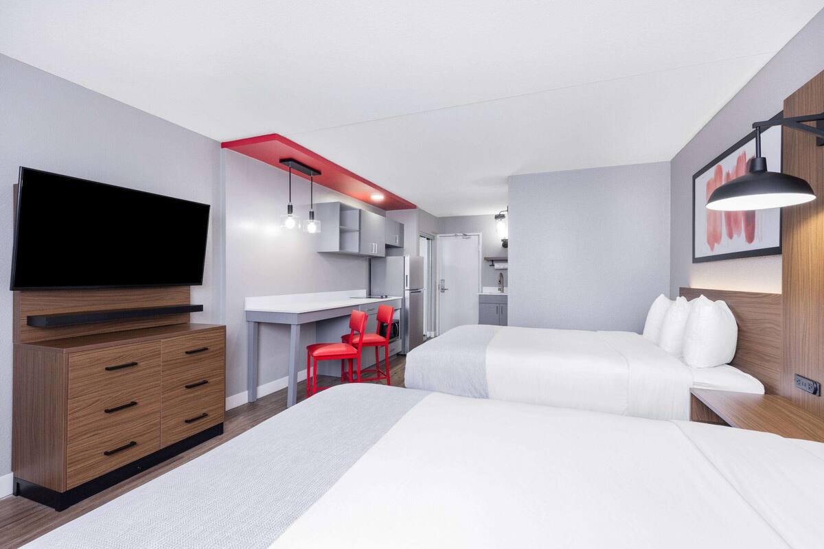 Una habitación de hotel moderna con dos camas, un televisor, un escritorio con una silla y una kitchenette.