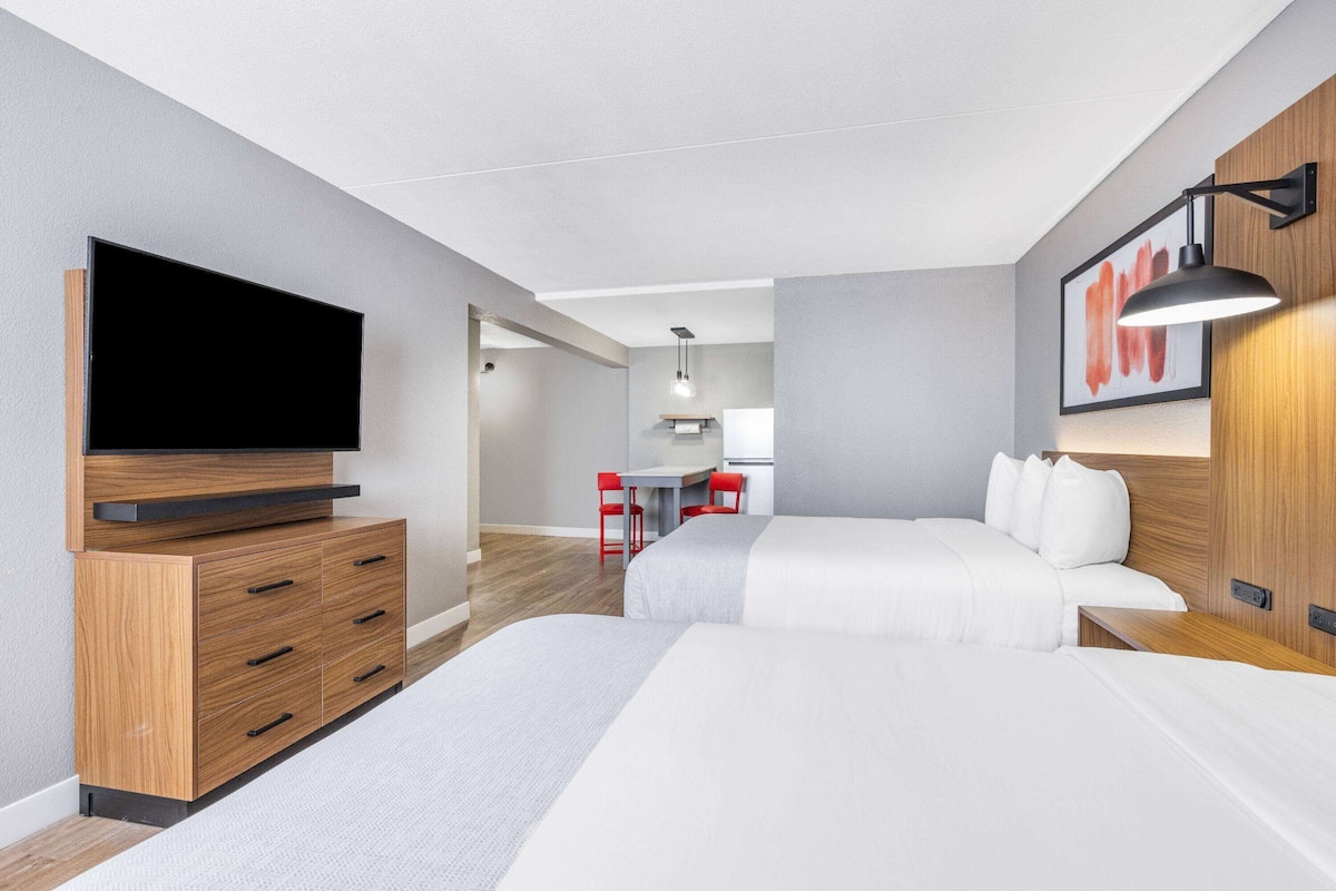 Una habitación de hotel moderna con televisión de pantalla plana, muebles de madera, un escritorio con sillas y una cama con ropa de cama blanca.