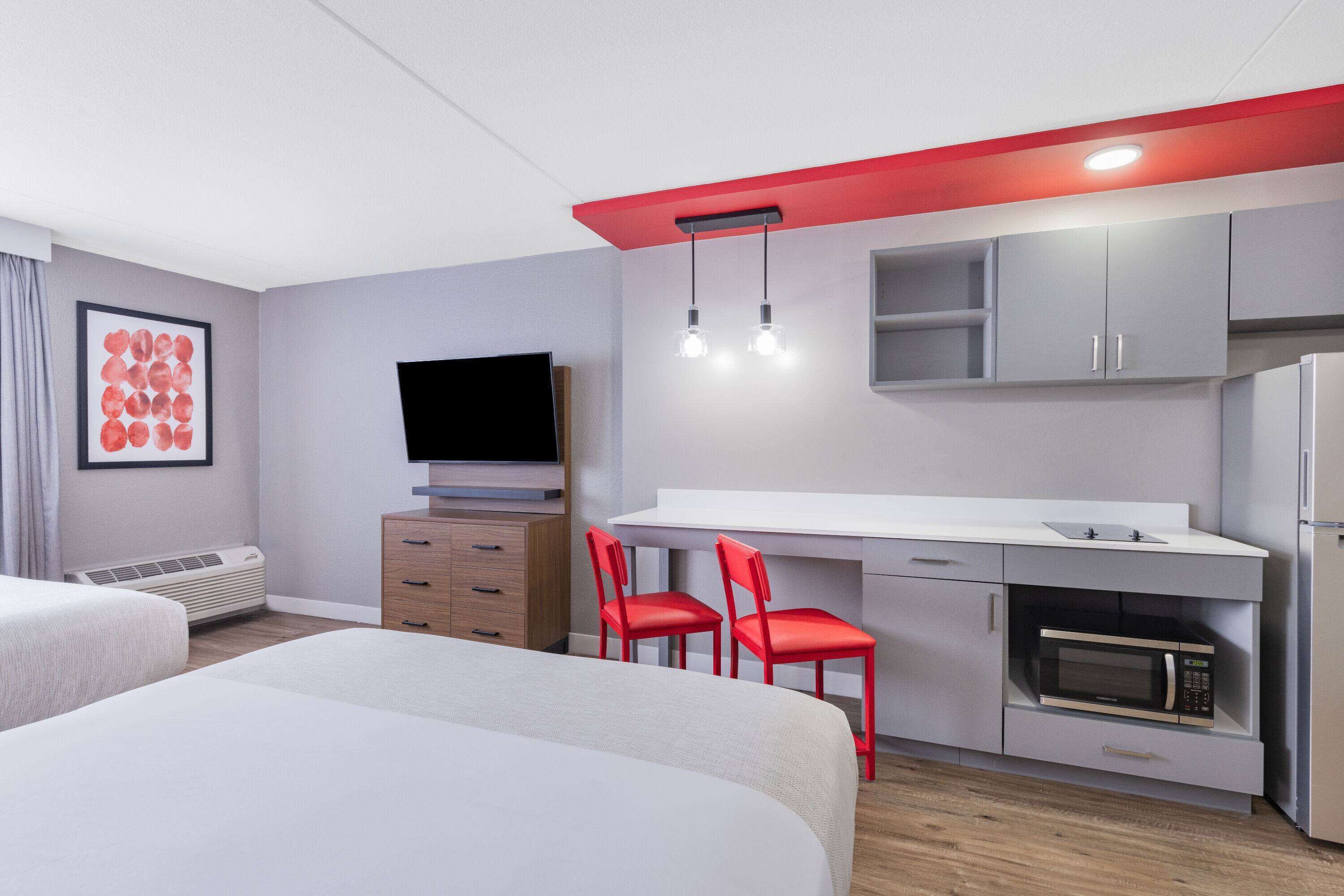 Una habitación de hotel moderna con una cama, un escritorio con dos sillas rojas, un televisor de pantalla plana, un microondas y una pequeña cocina.