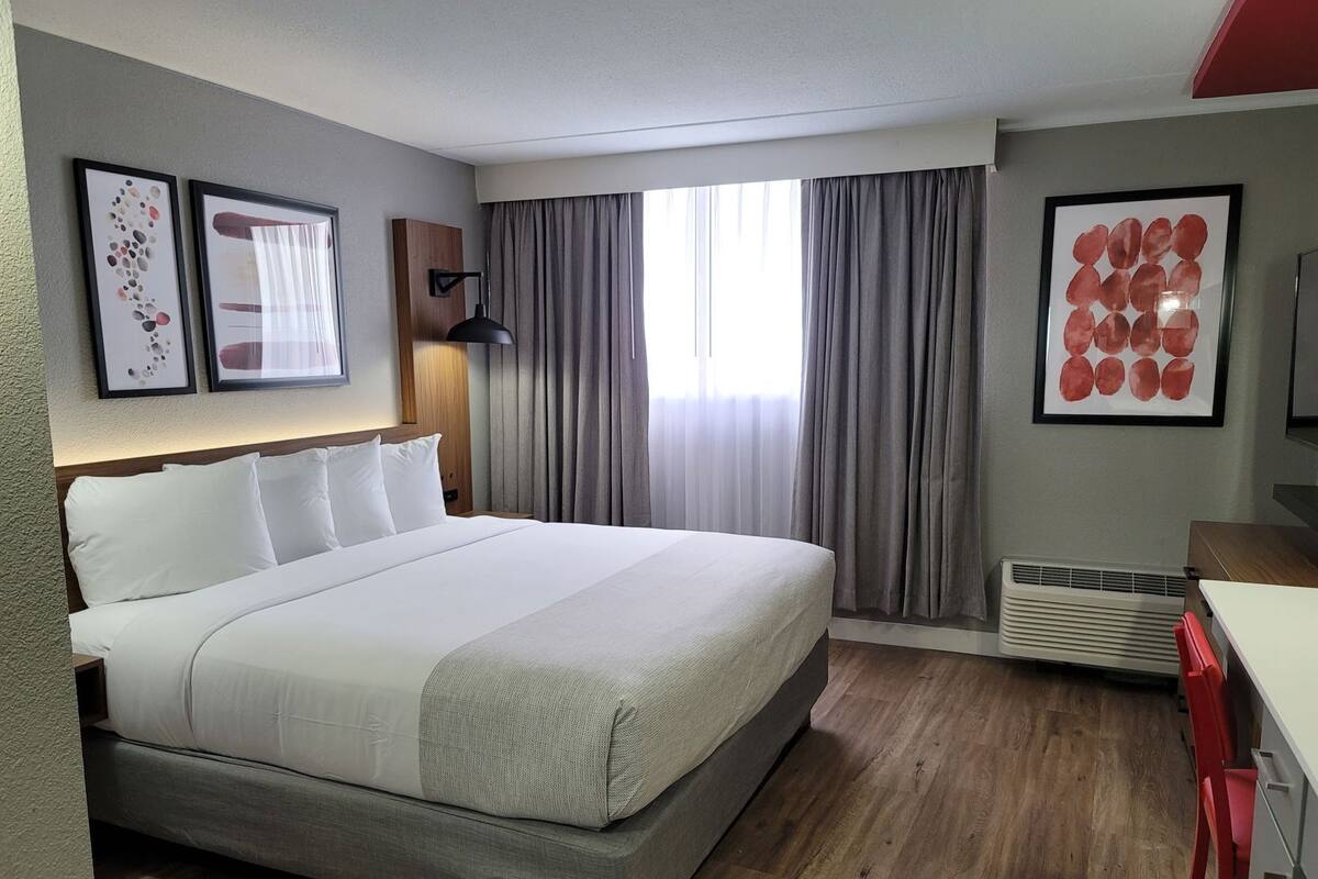 Habitación de hotel con una cama grande, un escritorio, una silla, una ventana con cortinas y obras de arte en las paredes.