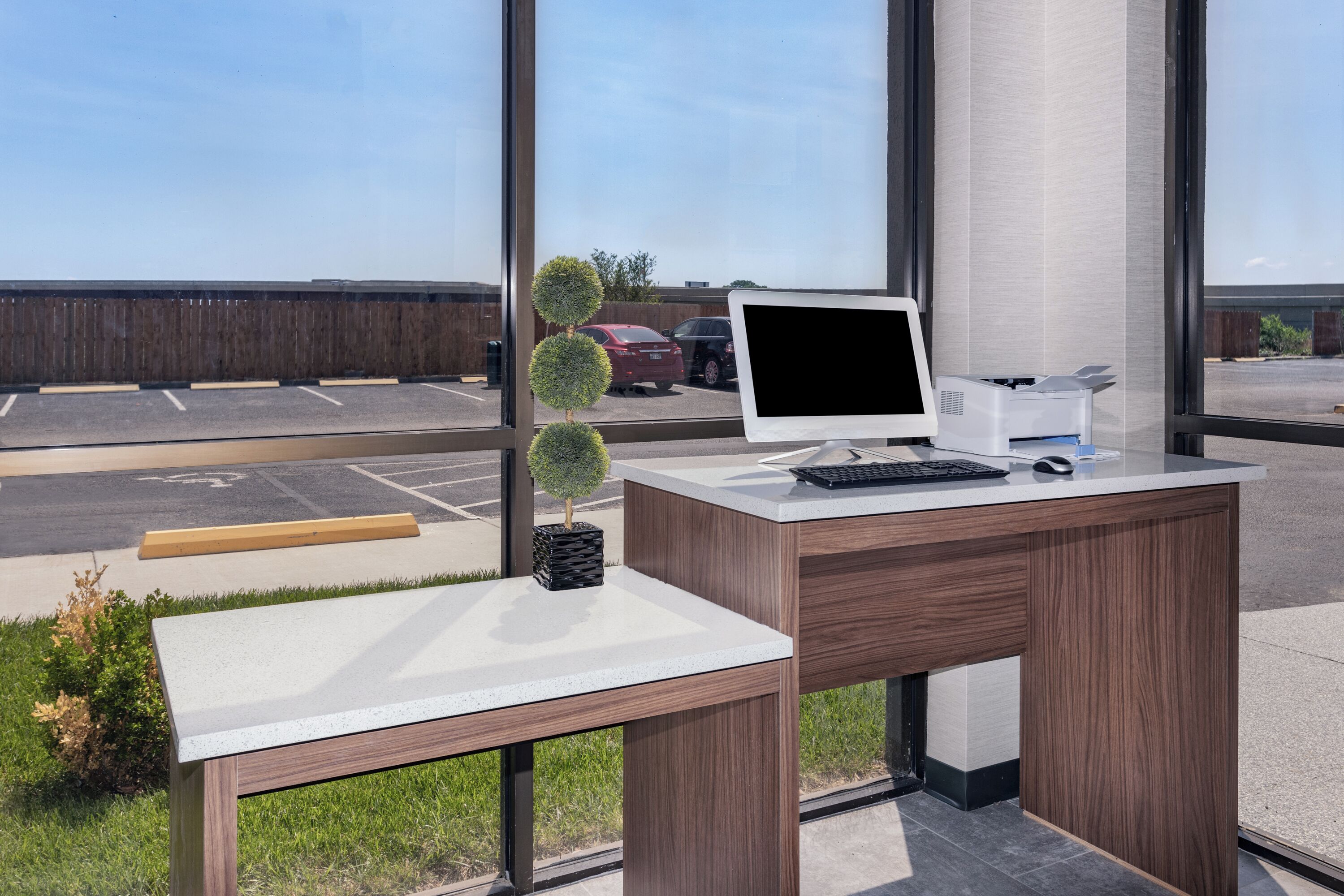 Un escritorio moderno de oficina con una computadora, teclado y ratón, ubicado cerca de amplios ventanales con vistas a un estacionamiento.