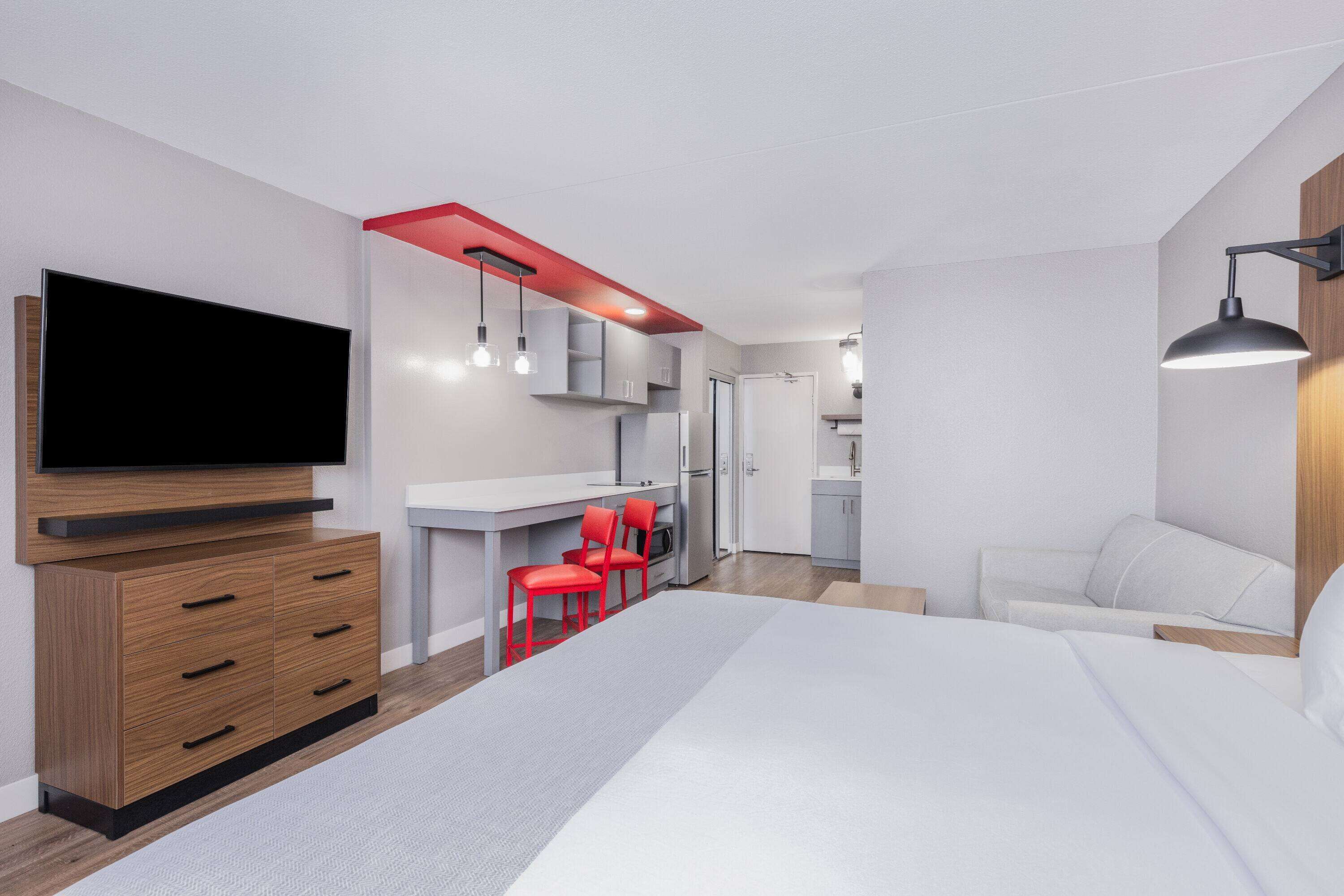 A modern bedroom with a large bed, a TV, a desk with red chairs, and a kitchenette.