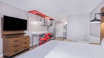 A modern bedroom with a large bed, a TV, a desk with red chairs, and a kitchenette.