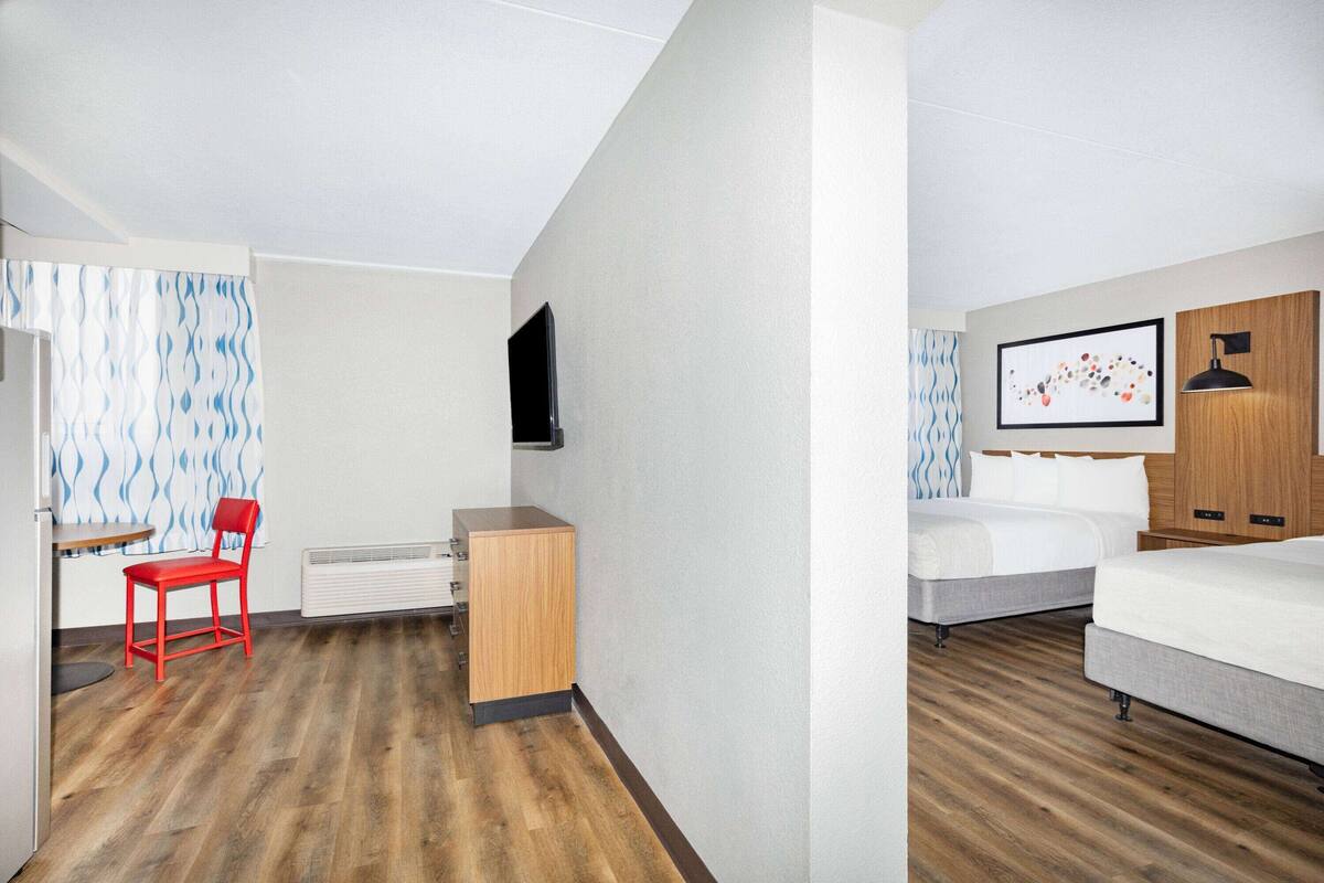 Habitación de hotel con piso de madera, una silla roja, un escritorio, un televisor, una cama y un cuadro en la pared.