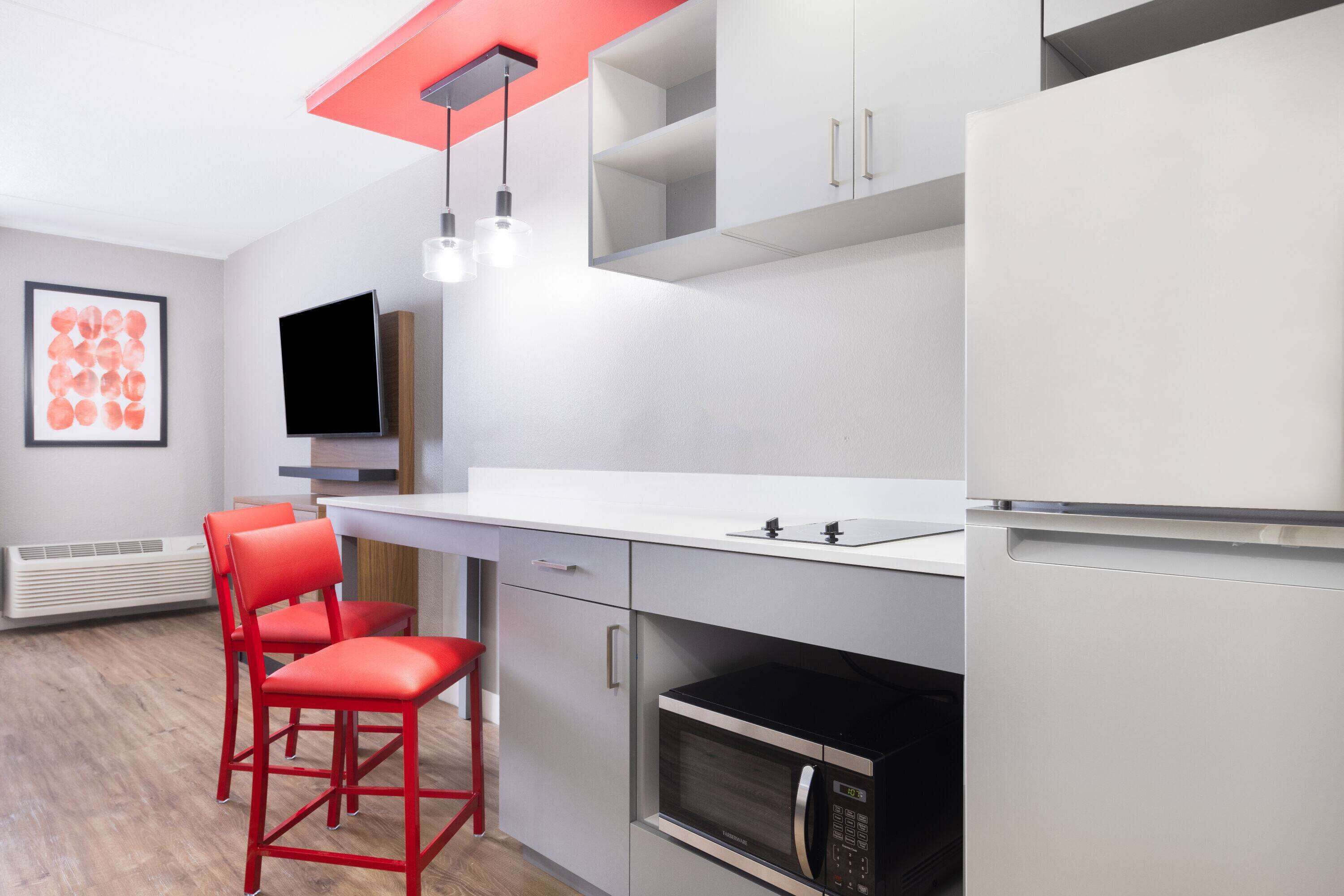 Una cocina moderna con encimera blanca, sillas rojas, un microondas y un televisor de pantalla plana.