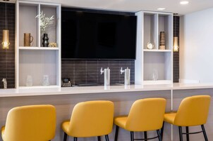 Una zona de bar moderna con una televisión de pantalla plana, estantes blancos y taburetes amarillos.