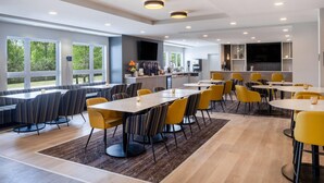 Un comedor moderno con varias mesas y sillas, grandes ventanales con vistas a zonas verdes y una cocina con televisor de pantalla plana.