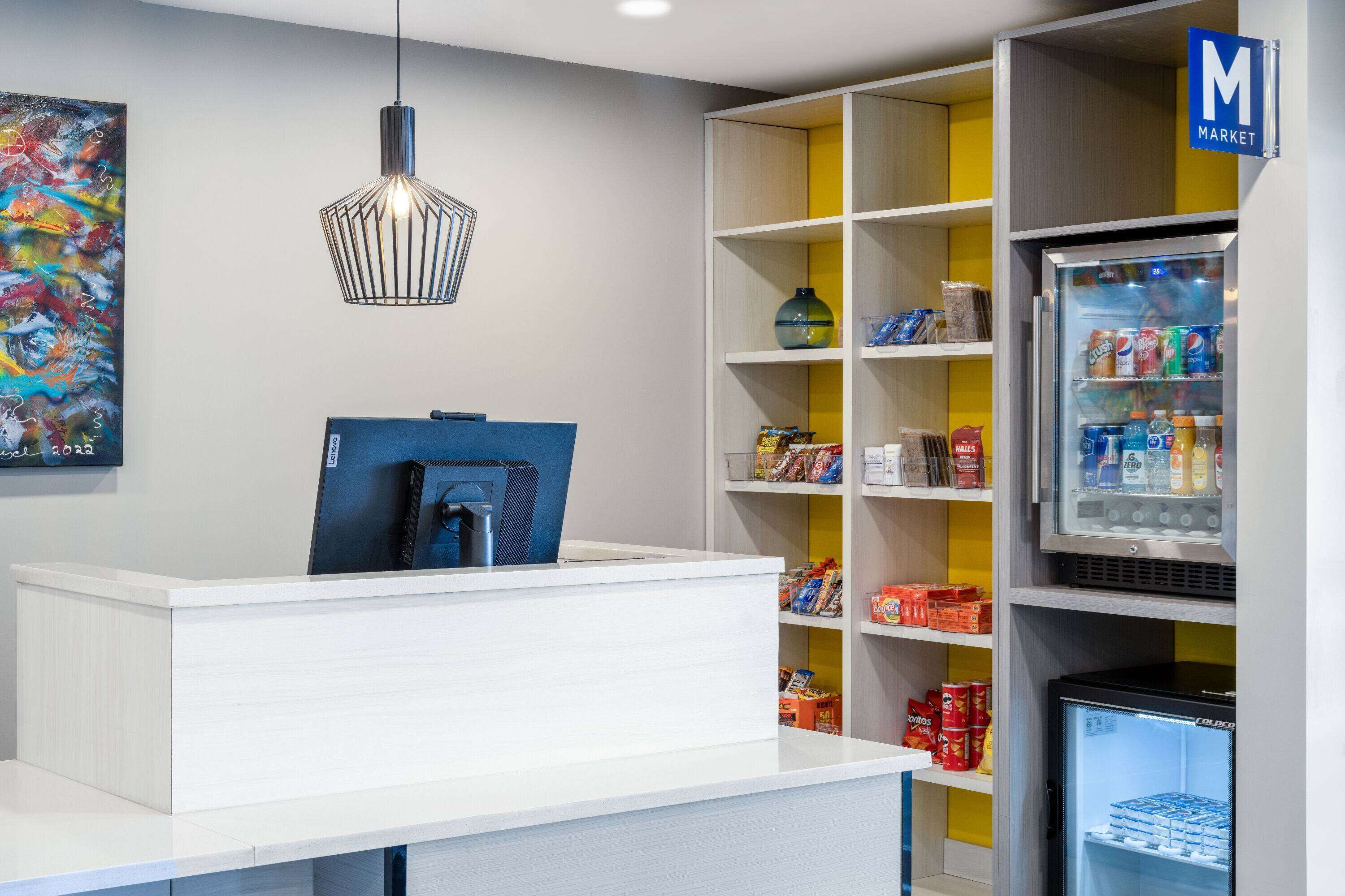 A reception area with a white counter, a blue chair, a hanging light fixture, a colorful abstract painting, and a vending machine labeled 'Market'.