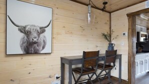 Interior de madera con zona de comedor que incluye una mesa y sillas, una obra de arte colgada de un toro, y una cocina visible a través de un arco.