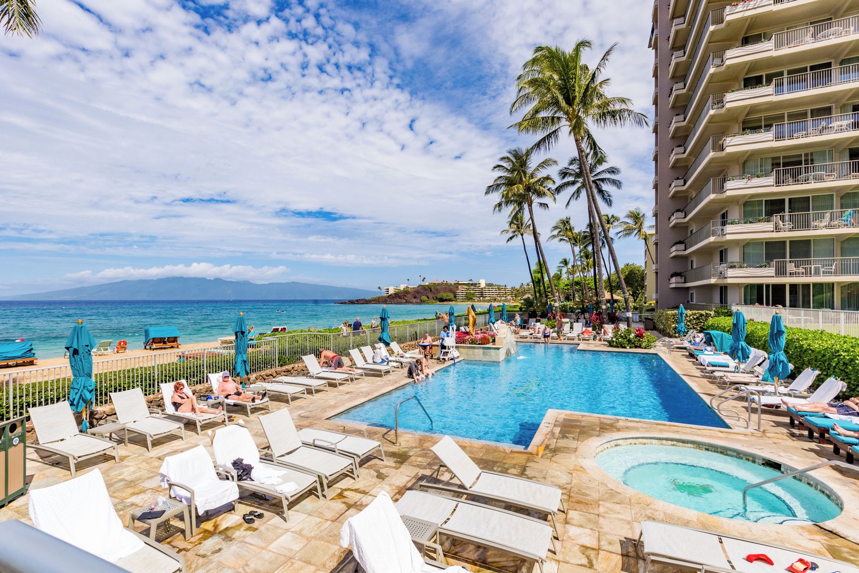 A resort pool area with multiple lounge chairs and umbrellas, a large central pool, and a view of the ocean.