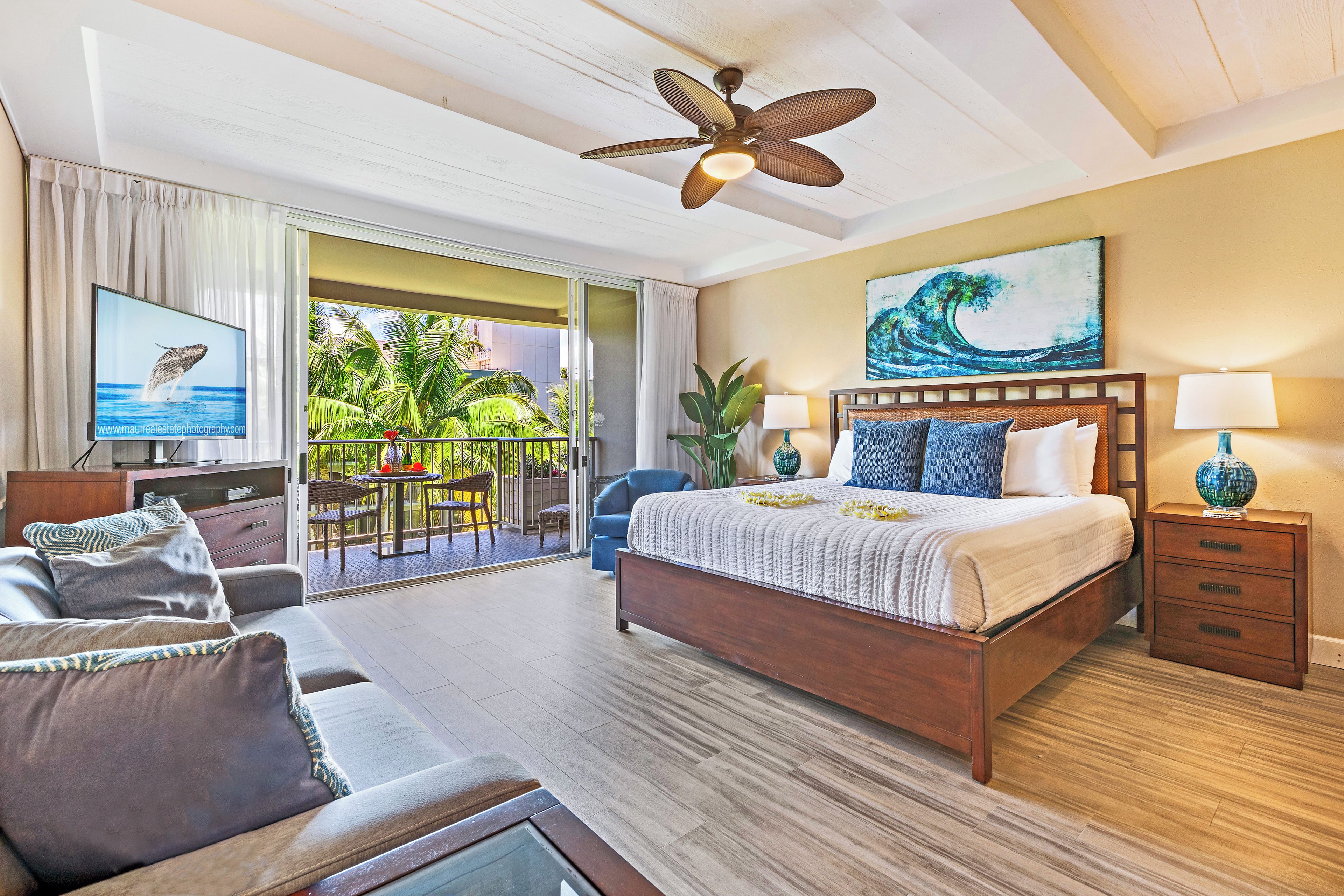 A hotel room with a bed, a TV, a balcony with a view of palm trees, and a ceiling fan.