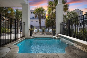 Un área de piscina con vista a un atardecer, rodeada de palmeras y un edificio blanco al fondo.