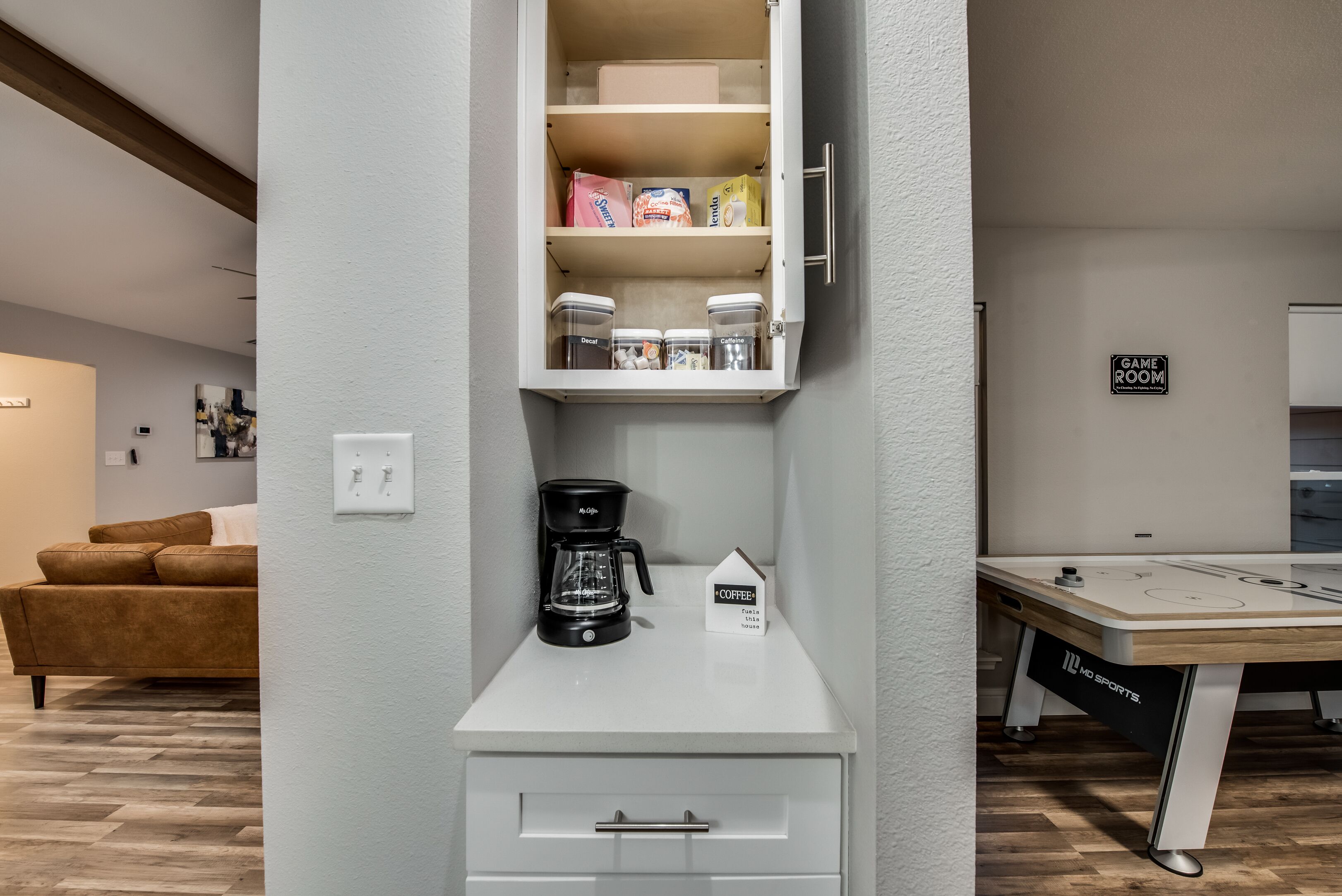 A kitchenette with a coffee maker, a cabinet with storage containers, and a small table with a coffee-themed sign.