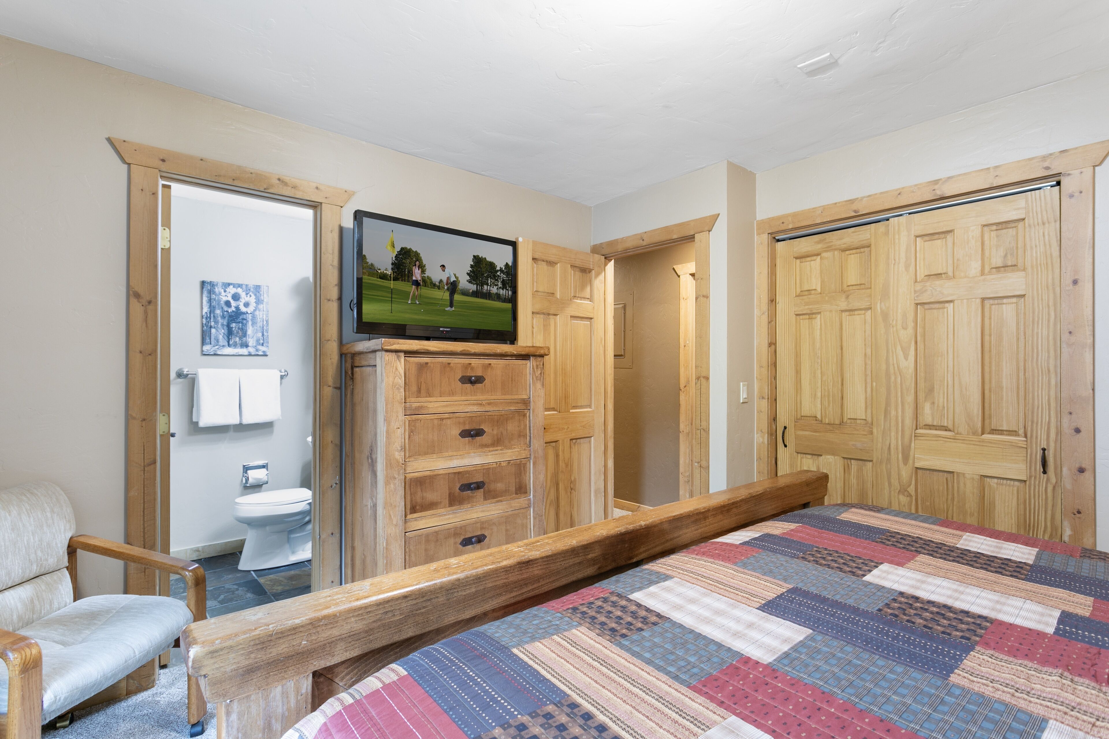 Un dormitorio con un marco de cama de madera, colcha, cómoda de madera, televisor y puerta de madera.